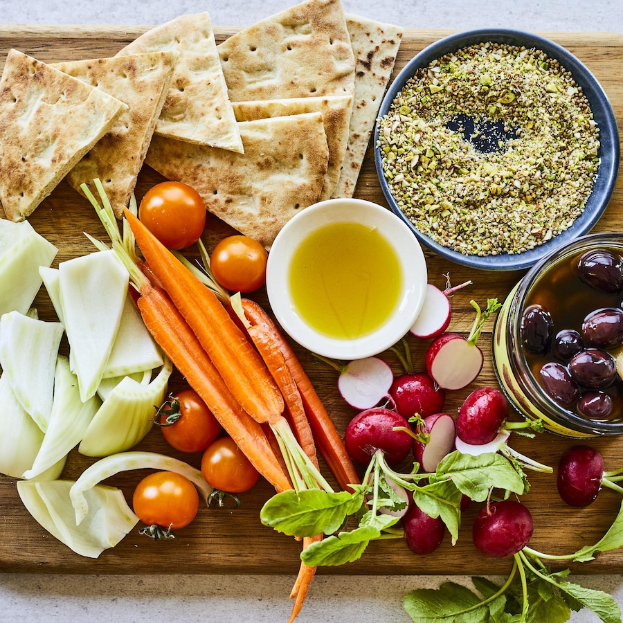 Sur une planche en bois : des carottes pelées, des olives Kalamata, des radis, des pitas grillés et des branches de fenouil forment un cercle autour d'un petit bol d'huile d'olive. 
