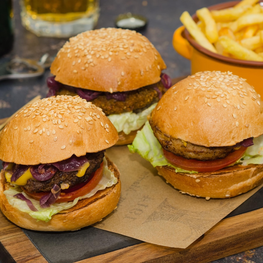 Trois burgers sur une planche en bois avec un bol de frites en arrière-plan.