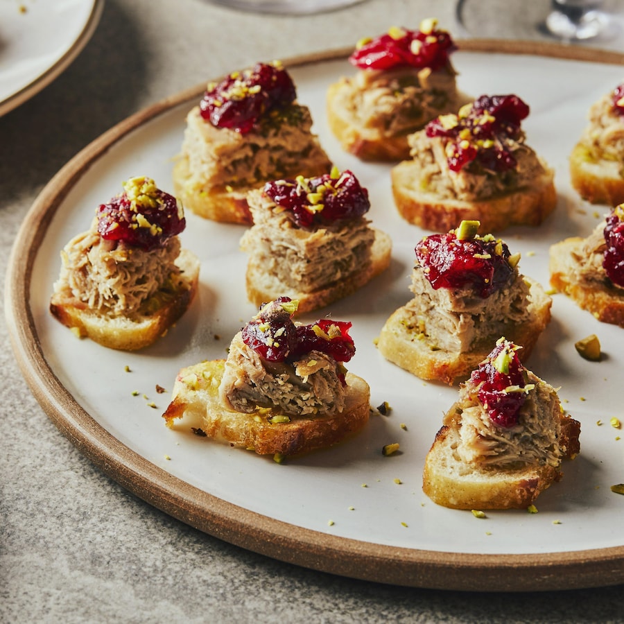 Dans une grande assiette, il y a une douzaine de canapés composés d’une tranche de baguette, un cube de rillette, une compote de canneberges et des pistaches émiettées.