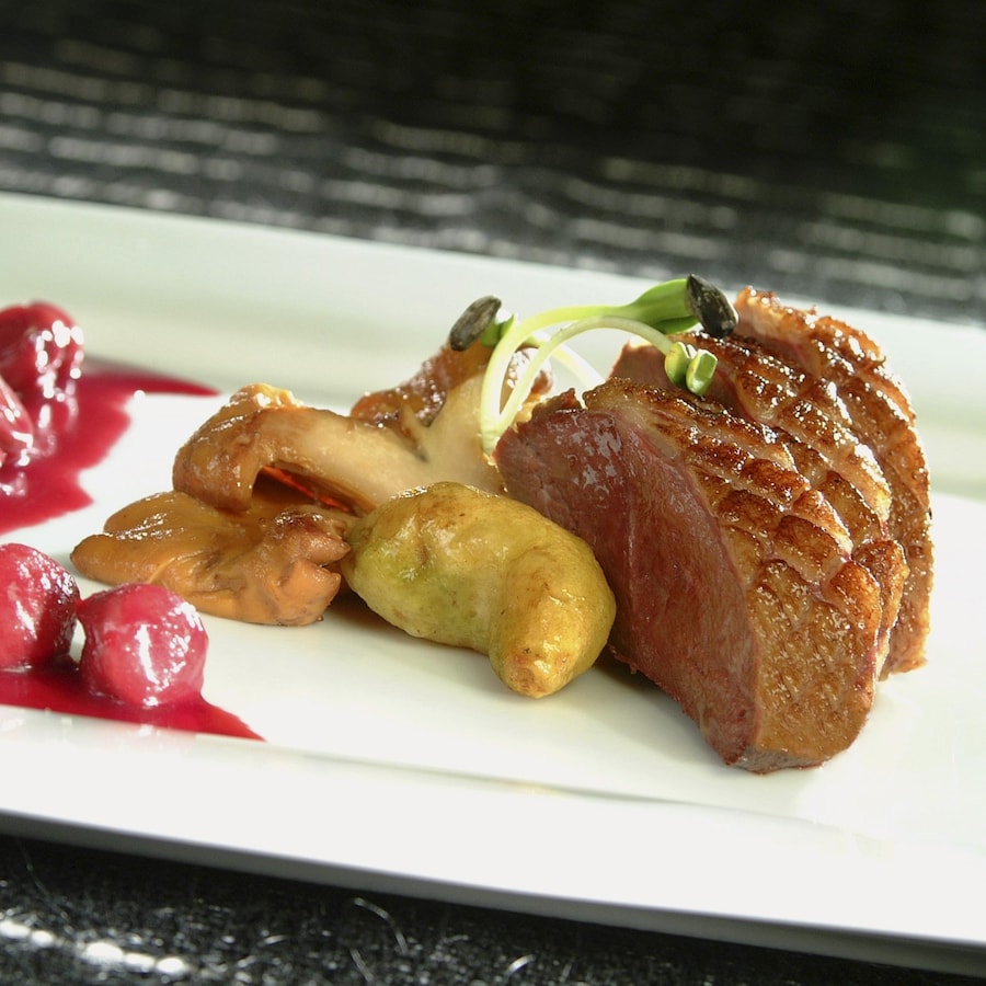 Trois tranches épaisses de canard Montmorency disposés dans une assiette blanche rectangulaire accompagnées de crosnes, de salsifis et de sauce rouge aux griottes.