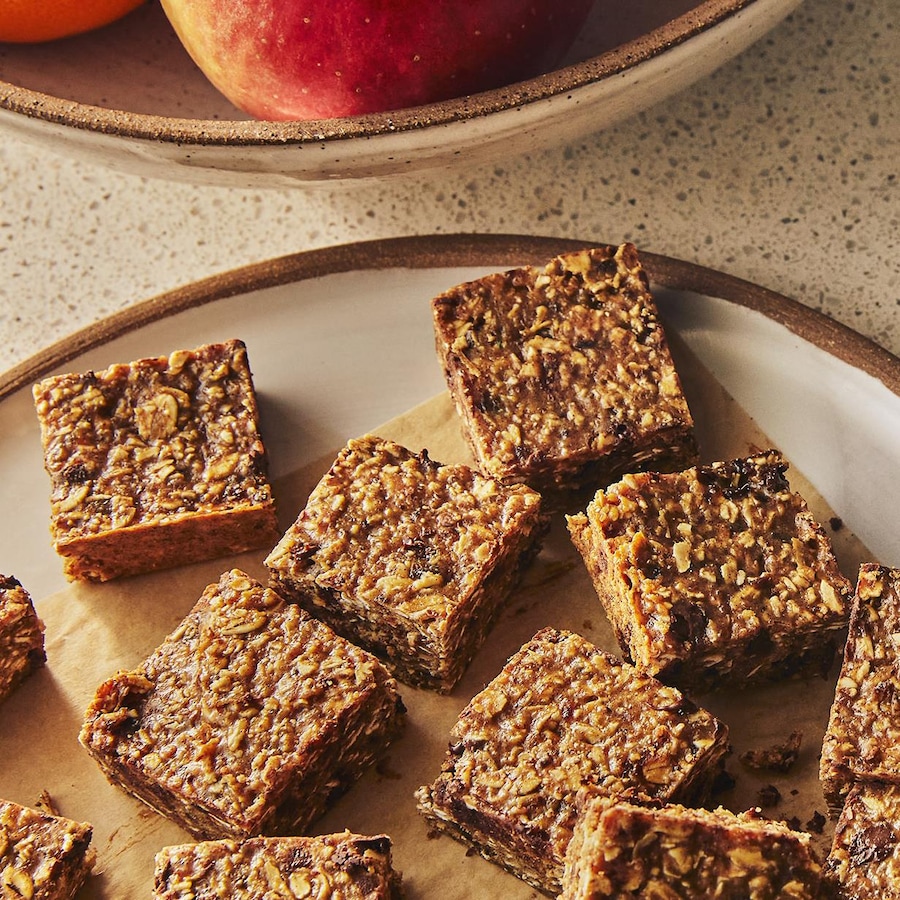 Des carrés protéinés au chocolat sont dans une assiette et dans un plat de conservation, à côté d’un bol de pommes.