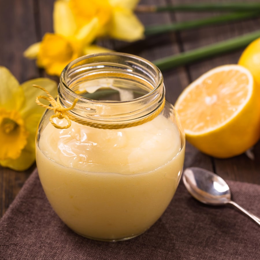 Un pot de crème au citron entouré d'une cuillère, de trois citrons et de deux jonquilles.