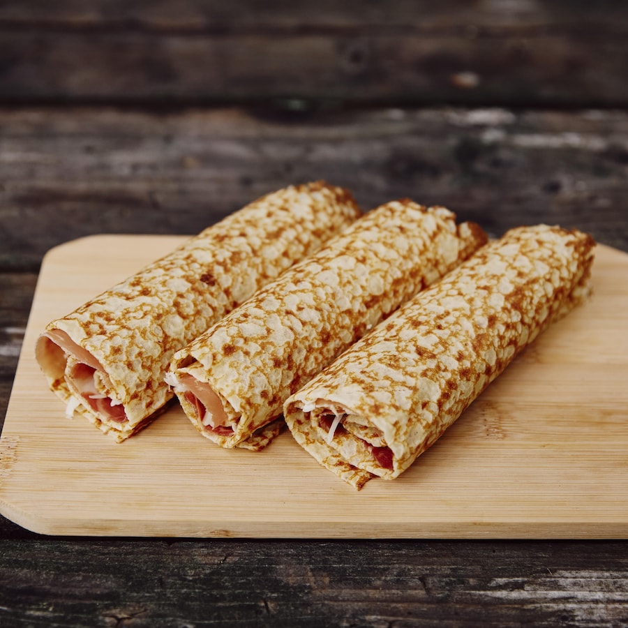 Une planche à découper en bois est déposé sur une table en bois. Sur cette planche à découper, il y a trois crêpes au fromage et au prosciutto.