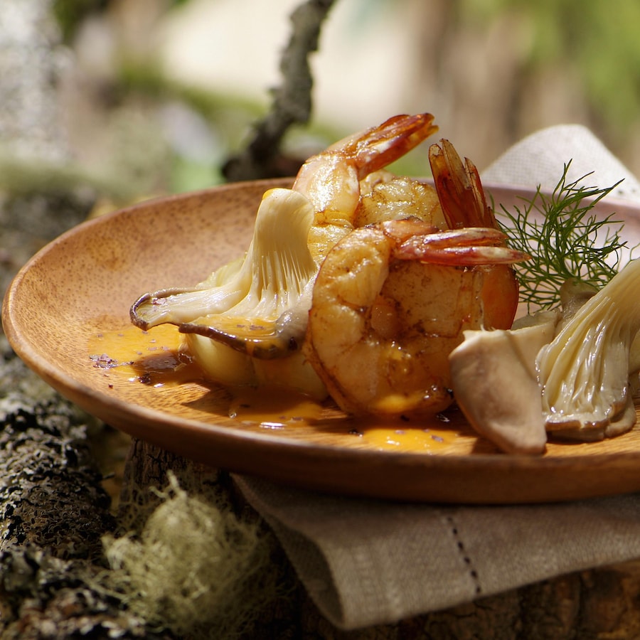 Deux crevettes cuites et deux pleurotes nappés d'une sauce légère orangée, et une branche d'anis, déposés dans une assiette brune.