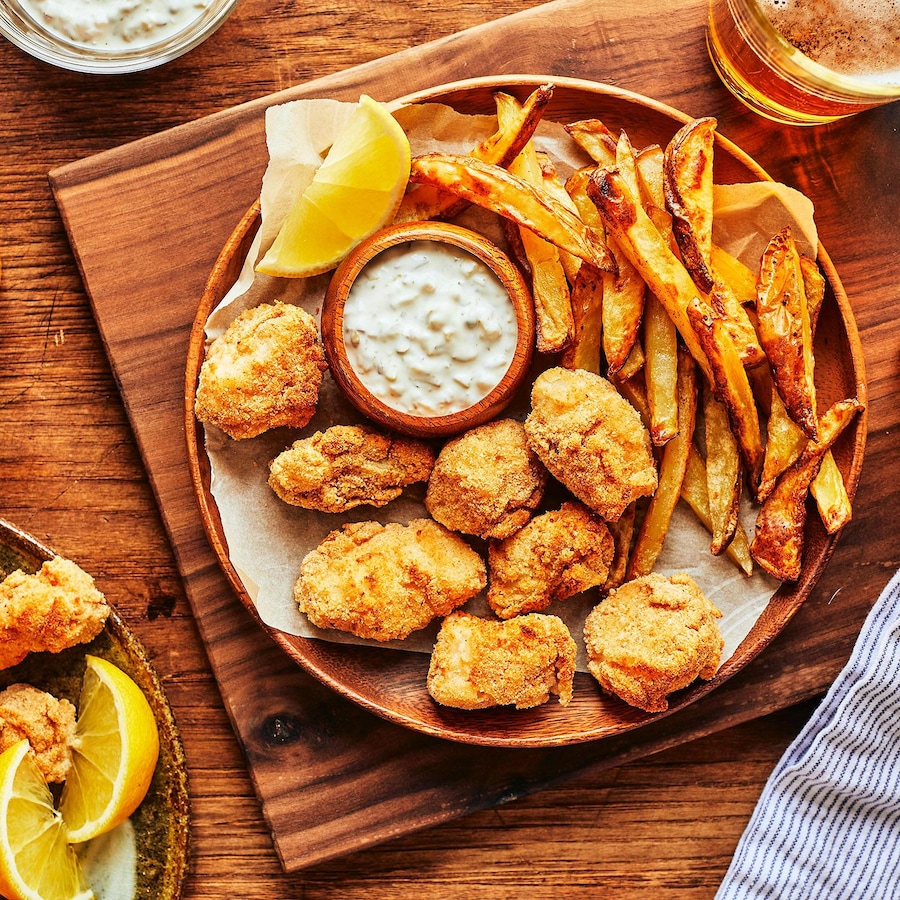 Des bouchées de fish and chips dans une assiette en bois sur une planche de bois, accompagnés de frites, d’une sauce tartare et de verres de bière. 