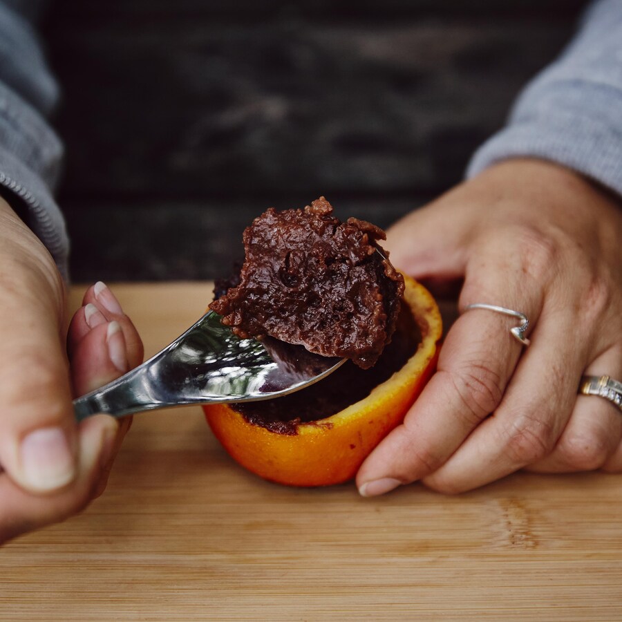 Sur une planche en bois, est déposé le gâteau au chocolat dans une orange.  Une des mains de Geneviève tient l'orange et l'autre tient une cuillère. Sur la cuillère, il y a un morceau de gâteau.