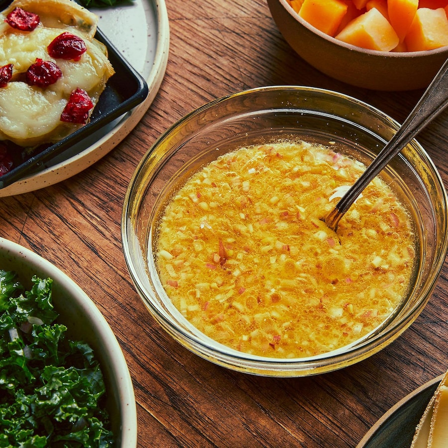 La marinade à raclette se trouve au centre d’une table, dans un bol en verre. Autour, il y a une assiette avec des patates gratinées et des canneberges cuites, un bol de kale, une assiette de fromage et un bol de rutabaga en cube.