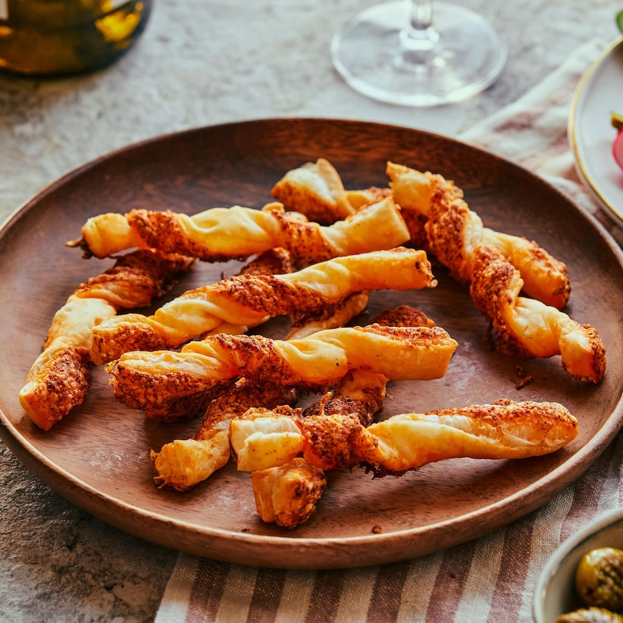 Des pailles feuilletées au fromage sont dans une assiette de bois avec un assiette de crudités, un pot d’olives et un verre de vin blanc.