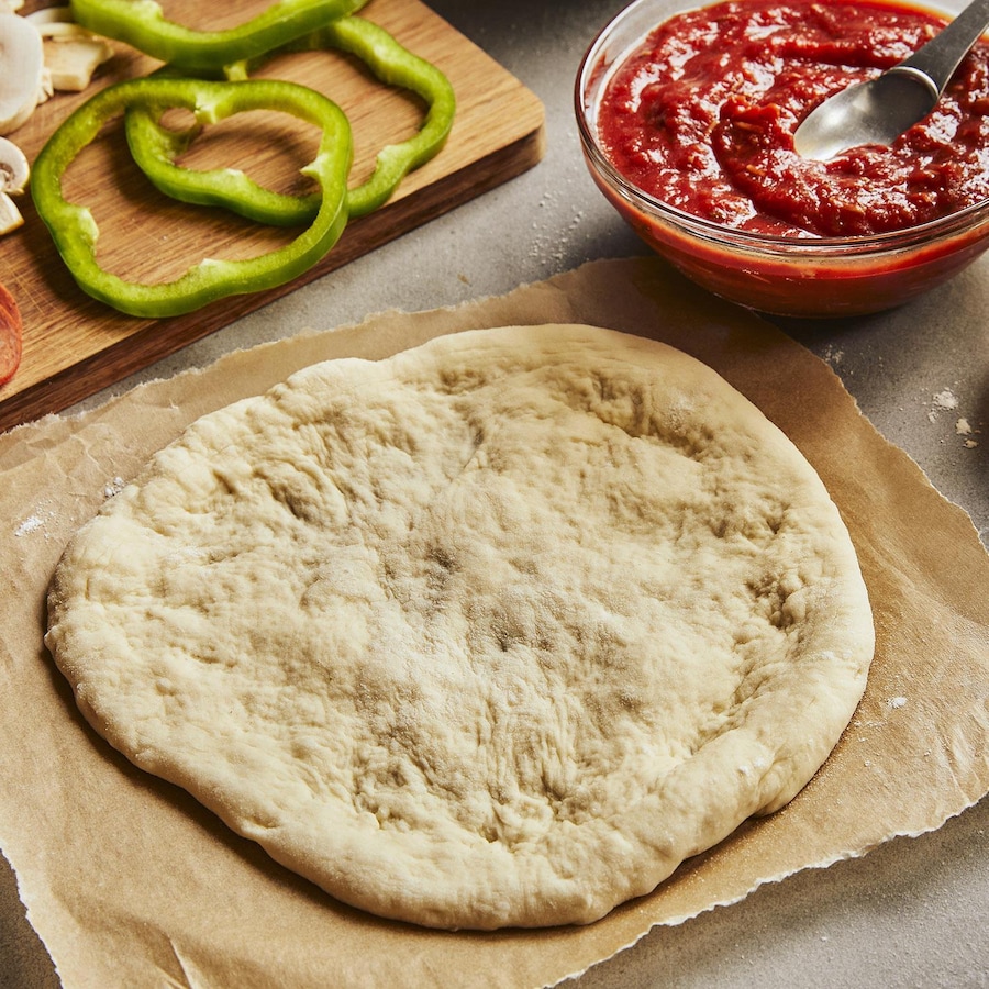 Une pâte à pizza déjà étirée est posée sur un papier parchemin. Autour, il y a une planche avec des légumes coupés et du pepperoni, un bol de sauce à pizza, un bol de fromage et un bol de farine.