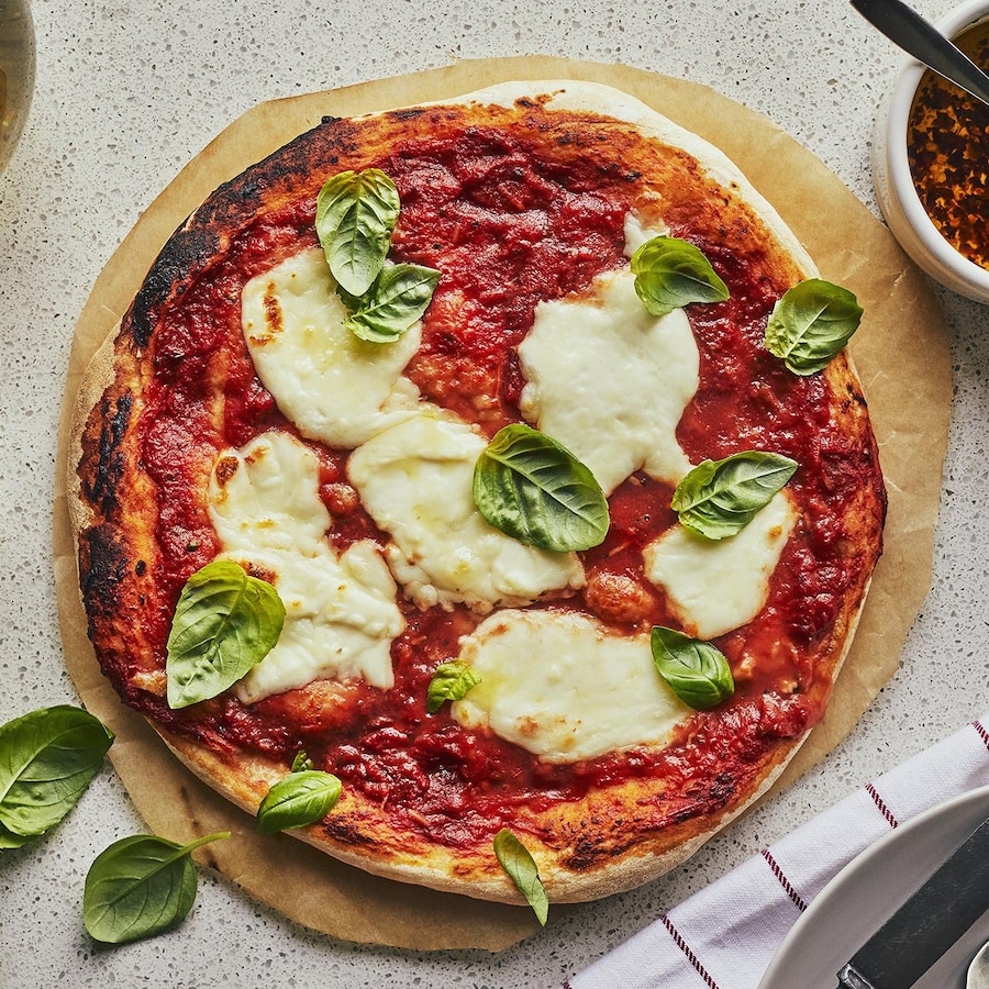 Au centre d’une table, une pizza Margherita est posée sur un papier parchemin rond. Autour, des feuille de basilic frais, une bouteille de vin blanc, un petit pot d’huile épicée et une assiette.
