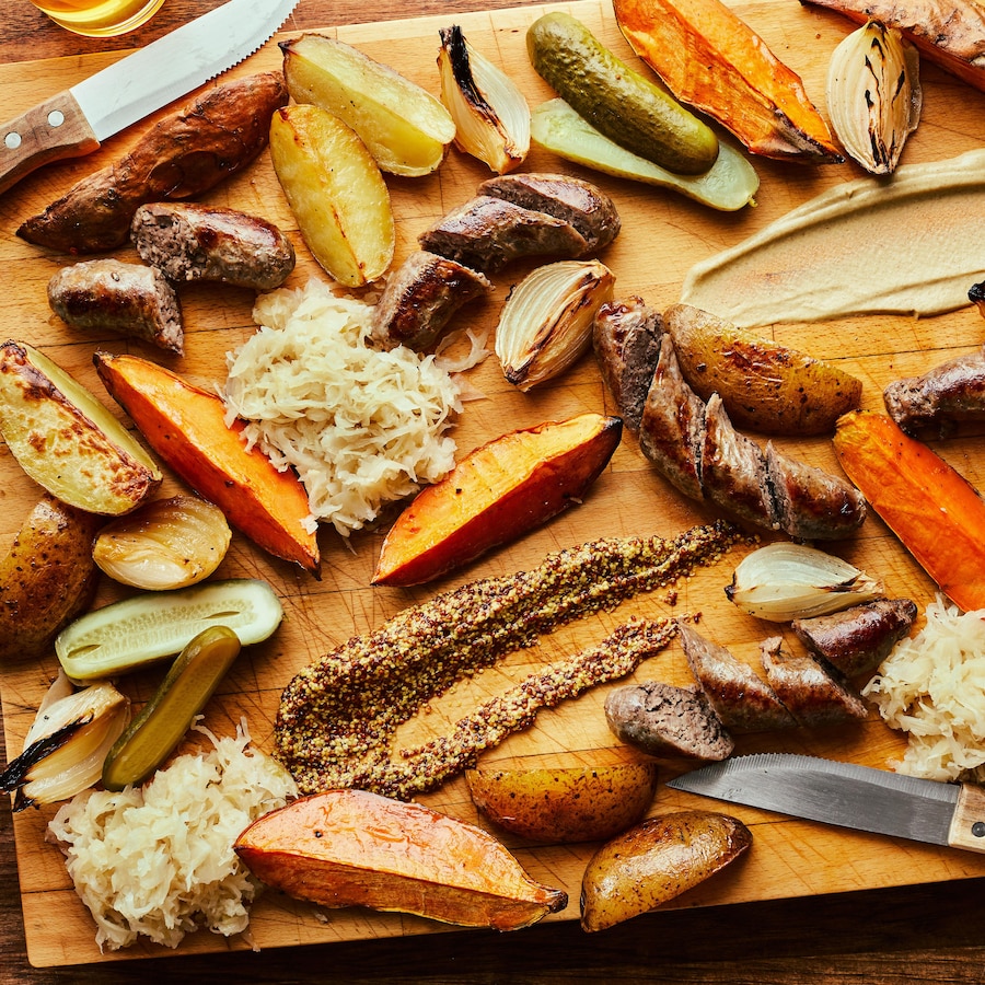 Il est possible de voir, sur une planche de bois, des saucisses, légumes rôtis, cornichons et choucroute.