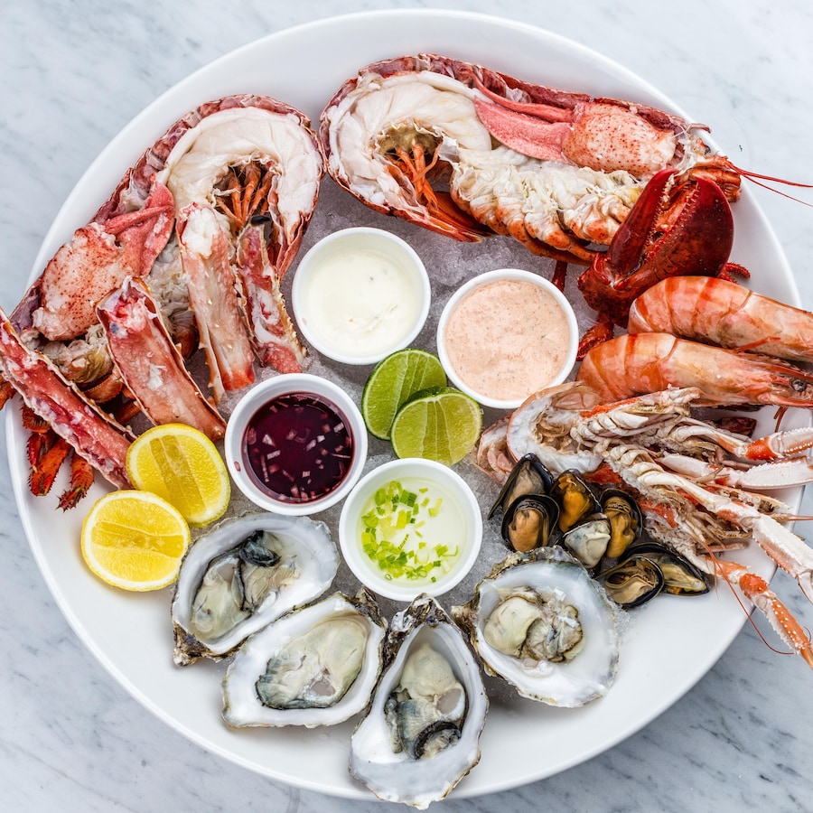 Un plateau avec du homard, du crabe, des huîtres, des moules et des crevettes servis sur glace, avec des sauces et des tranches d'agrumes.