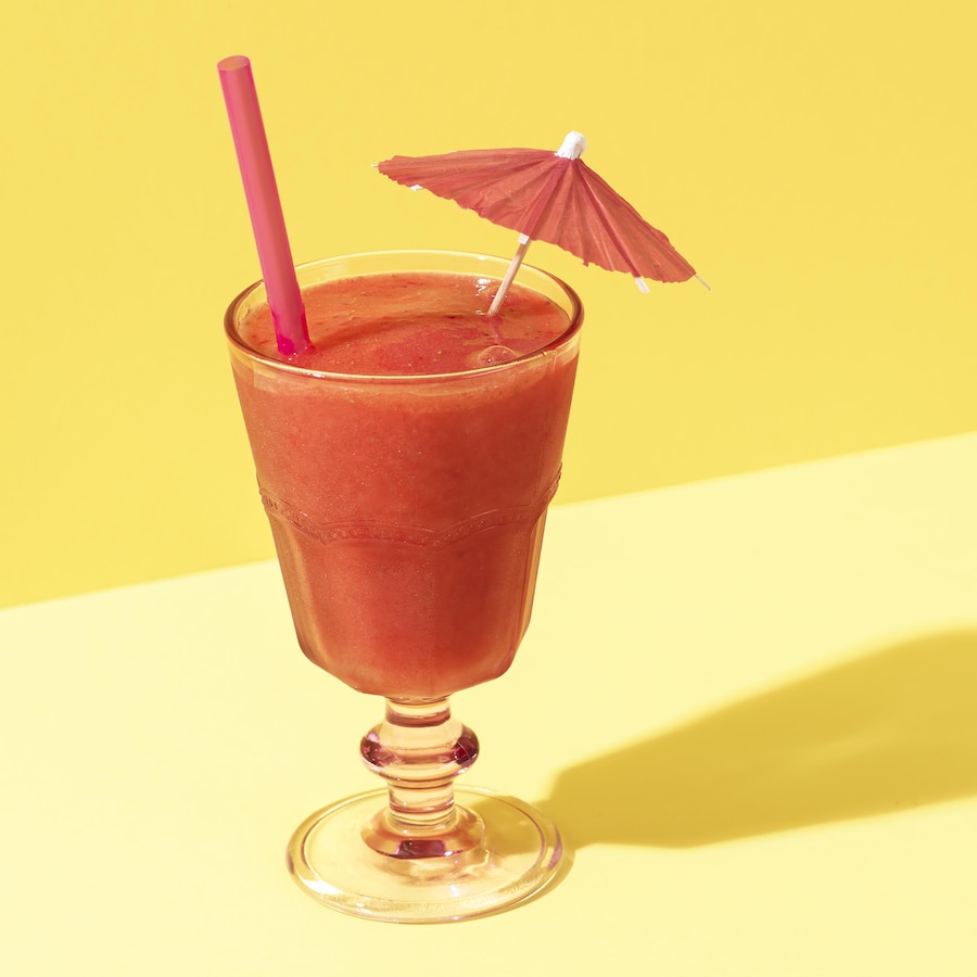 Un verre de liquide rose, paille, parasol de cocktail sur fond jaune.