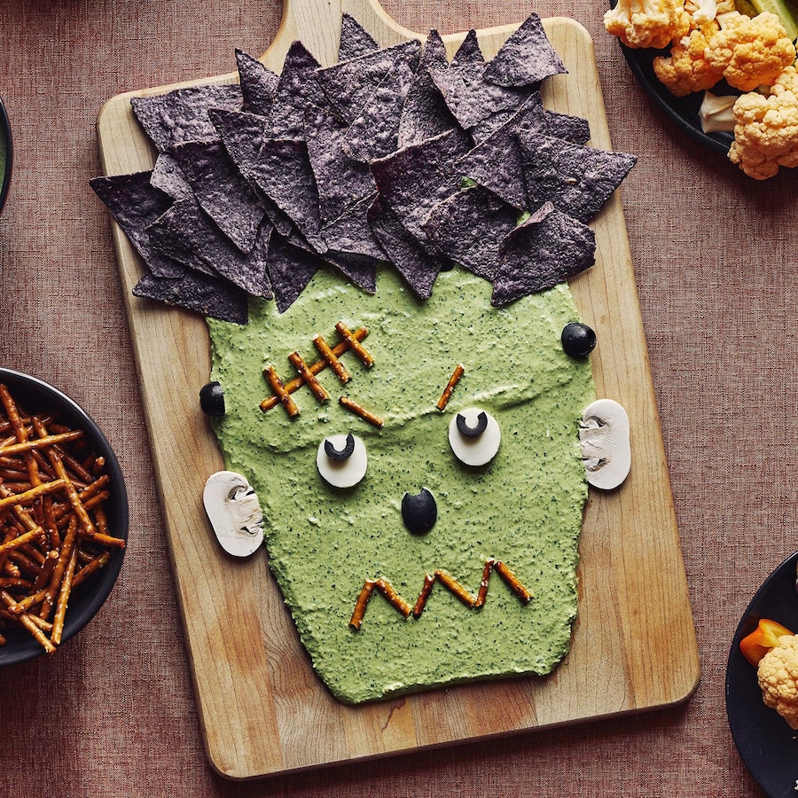 Une trempette en forme de visage de Frankenstein sur une planche de bois est accompagnée d’un bol de trempette, d’un bol de pretzels, d’un bol de croustilles de maïs bleu et de deux assiettes de légumes.