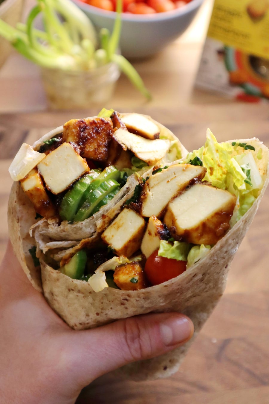 Une main tenant un wrap aux bâtonnets de tofu magique au BBQ et aux légumes, avec des ingrédients sur une table en bois en arrière-plan.