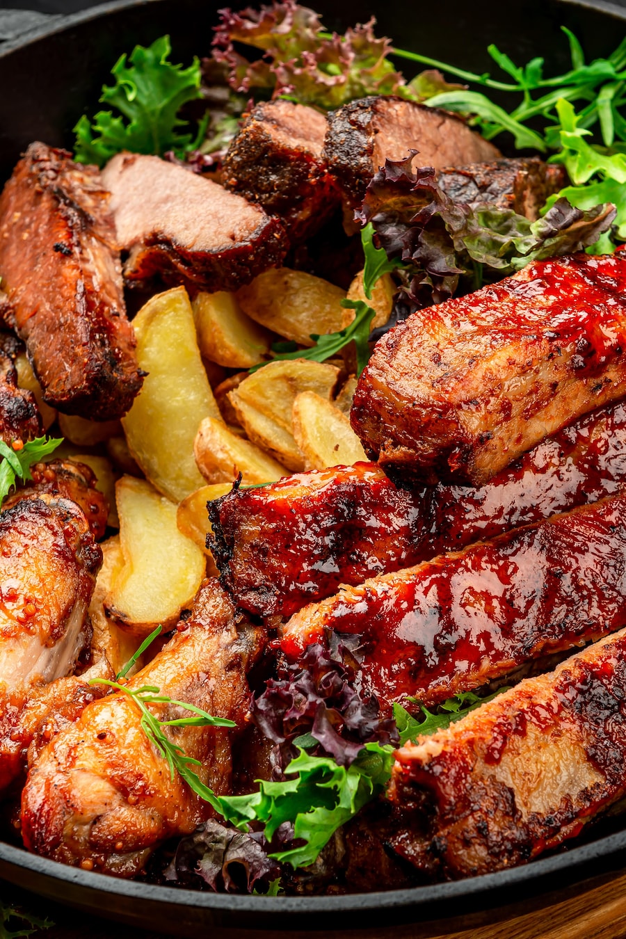 Un plat contenant des pommes de terres à la portugaise, des pilons de poulet et des morceaux d'agneau. La viande est couverte de sauce BBQ aux mangues.