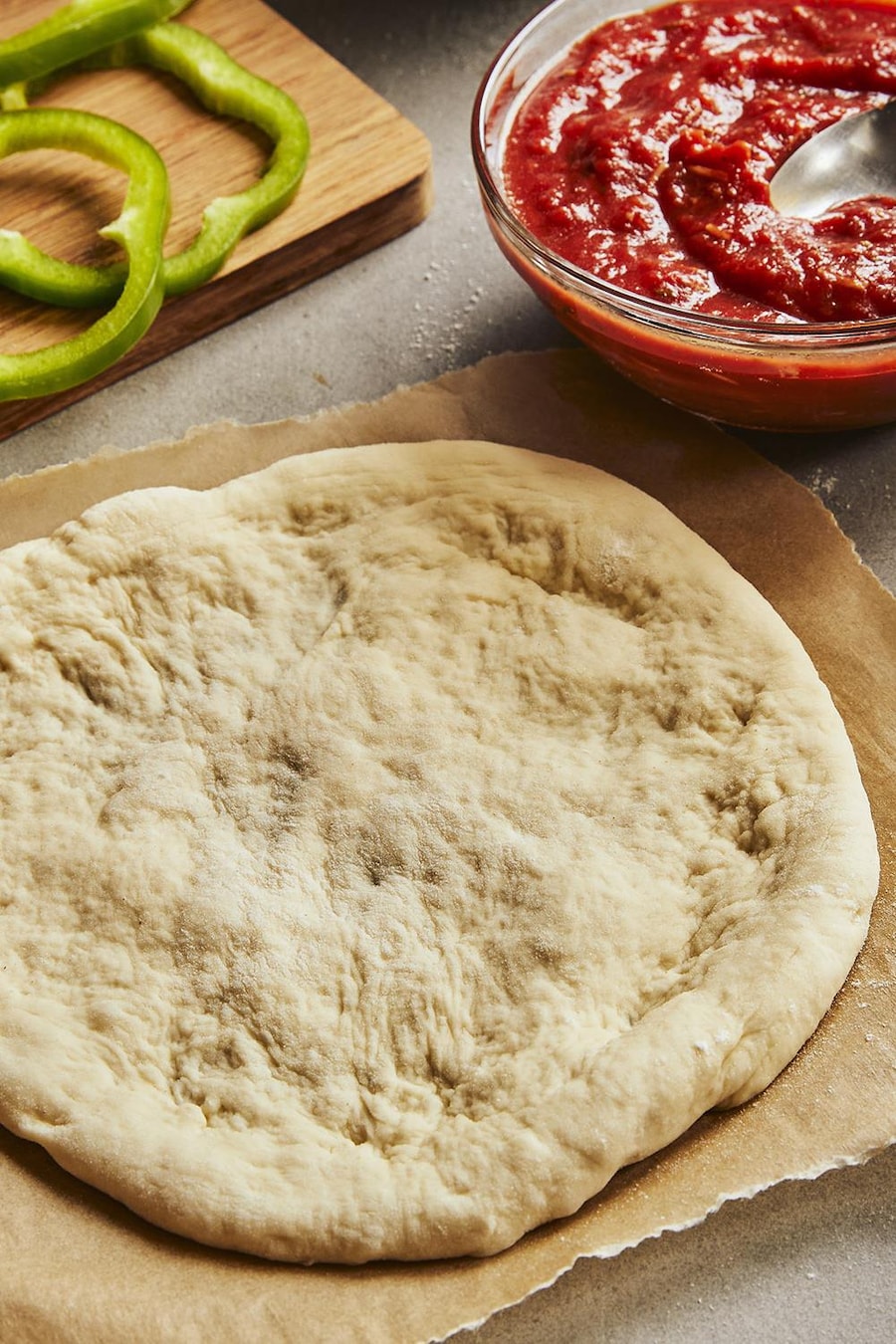 Une pâte à pizza déjà étirée est posée sur un papier parchemin. Autour, il y a une planche avec des légumes coupés et du pepperoni, un bol de sauce à pizza, un bol de fromage et un bol de farine.