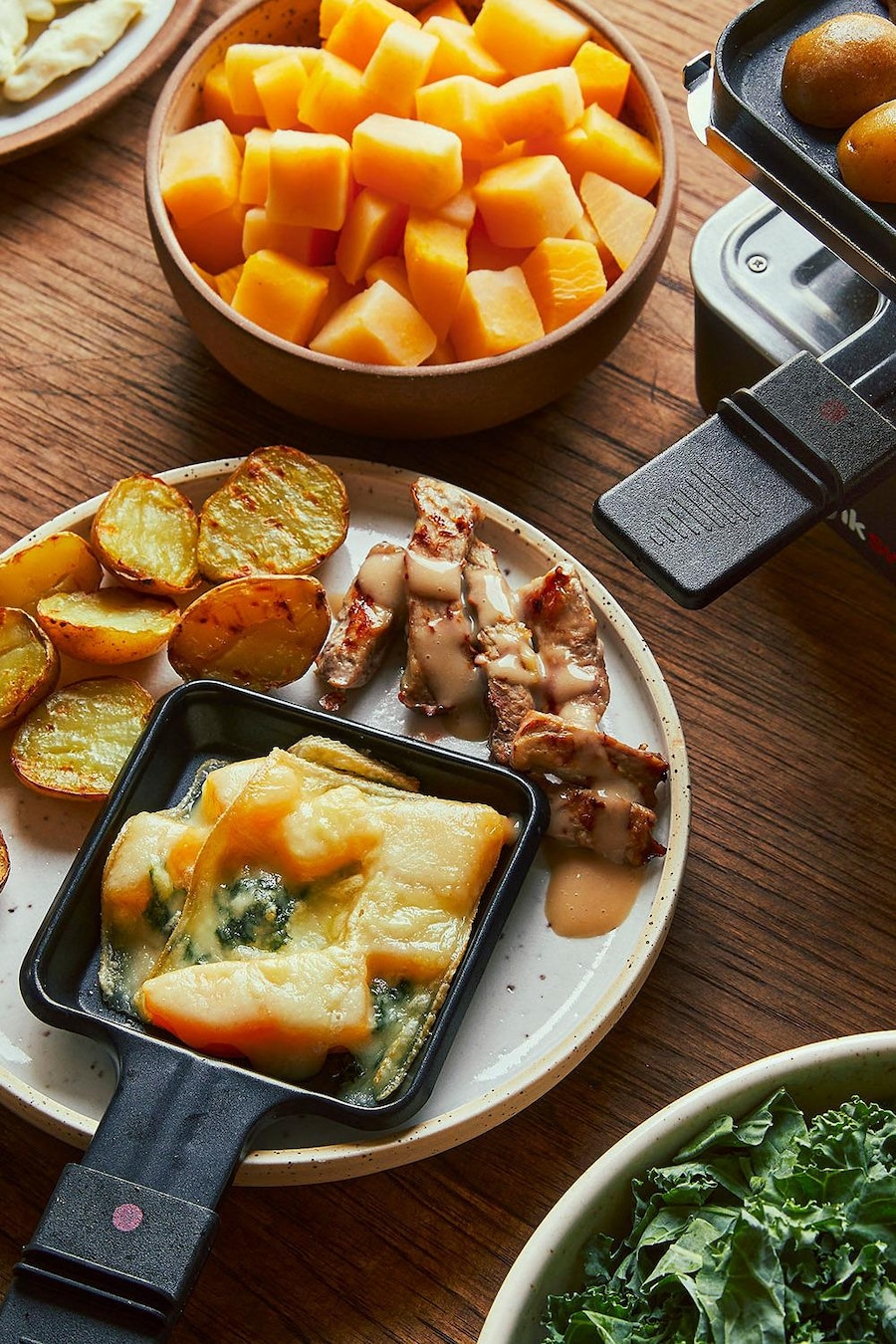 Une assiette avec des patates grillées, des petits morceaux de porc cuit et une coupelle à raclette remplie de kale et de rutabaga gratinés.