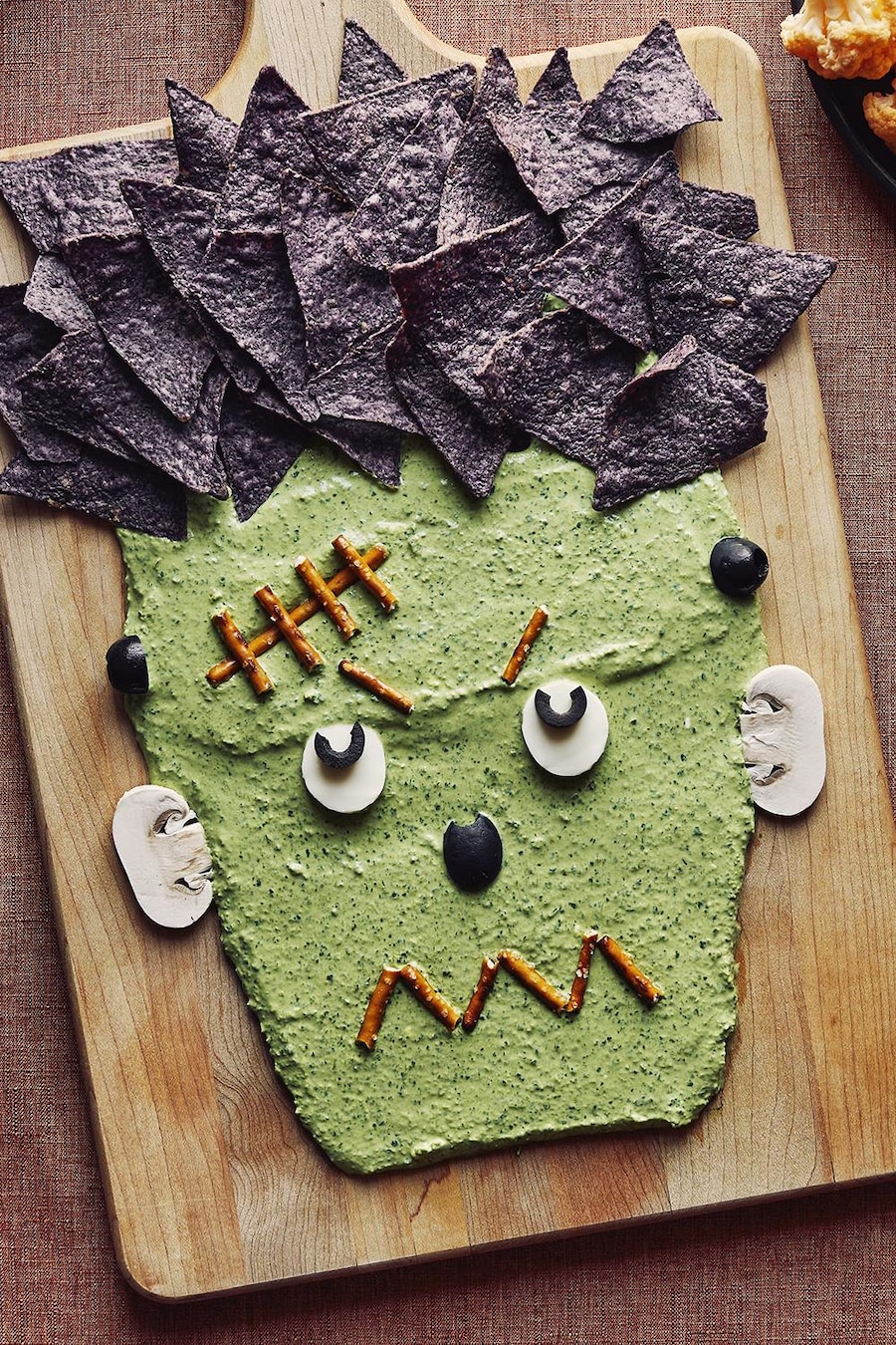 Une trempette en forme de visage de Frankenstein sur une planche de bois est accompagnée d’un bol de trempette, d’un bol de pretzels, d’un bol de croustilles de maïs bleu et de deux assiettes de légumes.