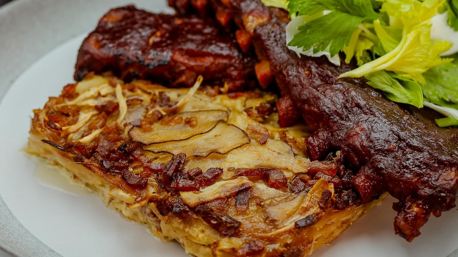 Un gratin de panais, pommes de terre rattes et bacon qui accompagne les côtes levées dans une assiette.