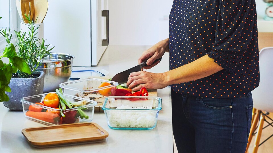 Une femme qui tranche des légumes sur un plan de travail avec différents plats réutilisables en verre.