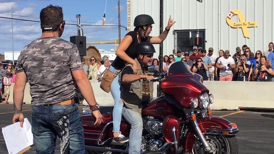 Un homme est assis sur un motocyclette rouge avec une femme comme passagère debout derrière. Elle salut la foule de gens qui observe l'événement.