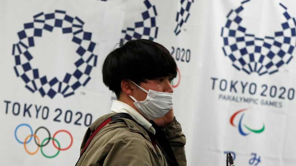 Un homme d'origine asiatique porte un masque sanitaire. Derrière lui, un bandeau fait la promotion des Jeux olympiques de Tokyo.