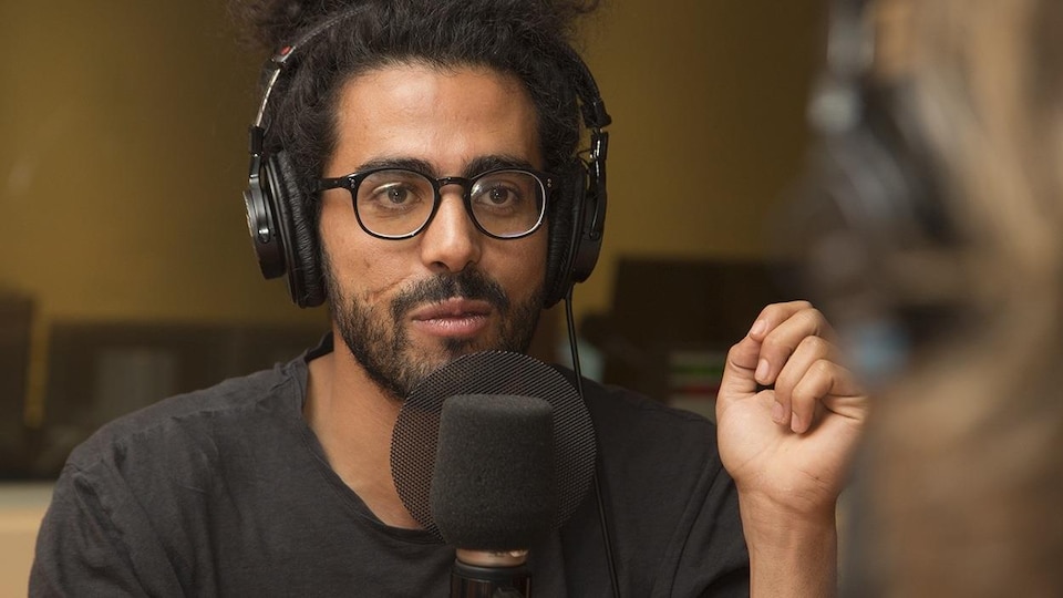 Portrait d'Adib Alkhalidey au micro d'une émission de radio. Il porte des lunettes noires et une barbe.