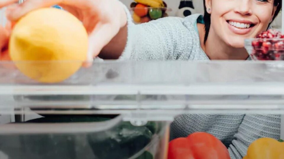 Vue de l'intérieur d'un réfrigérateur où l'on voit des aliments et le visage du'une femme souriante.