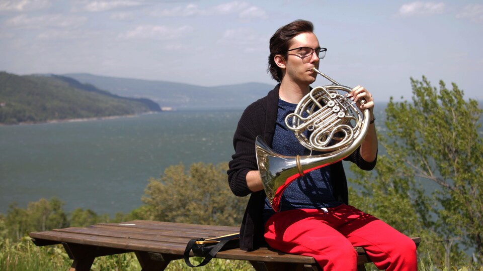 Un jeune homme joue du cor français, assis sur une table en bois, derrière lui, se trouvent le fleuve et les montagne de Charlevoix.