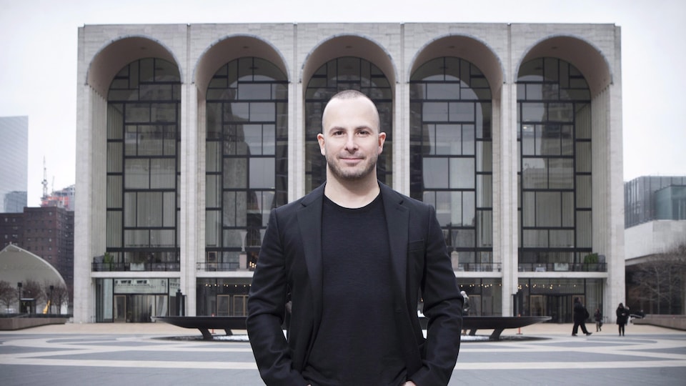 Le chef d'orchestre esquisse un sourire et pose à l'extérieur devant un grand bâtiment vitré.