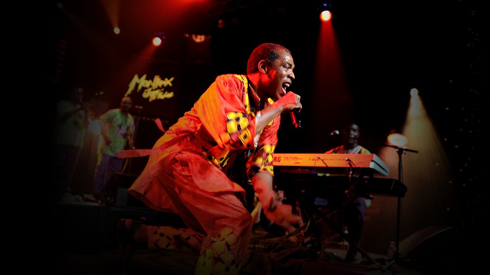 A man wearing an orange dress sings on stage holding a microphone. 