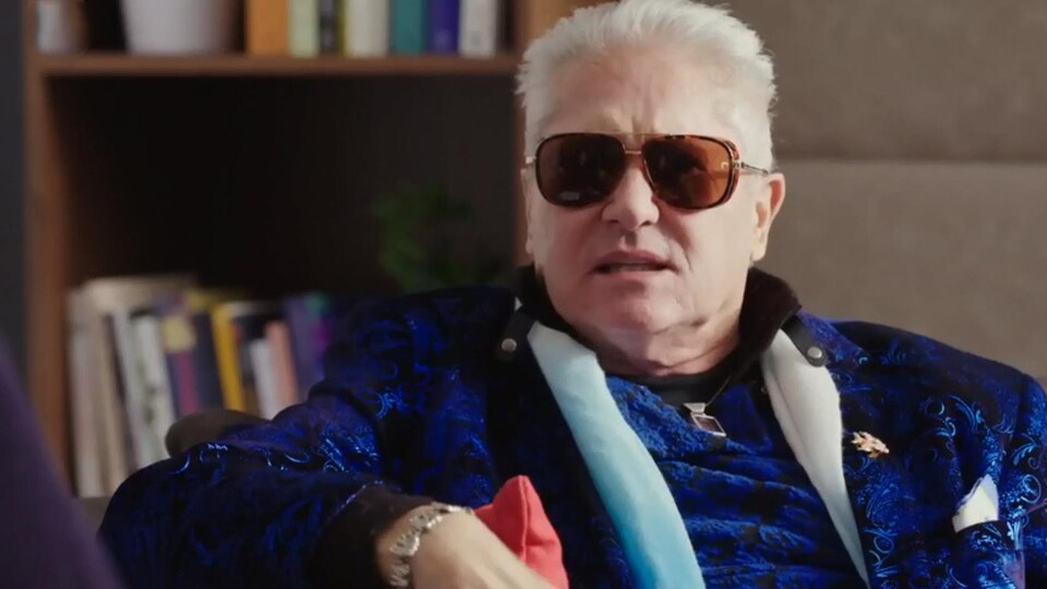 Le poète québécois Jean-Paul Daoust est assis ; il porte un veston bleu et des lunettes fumées.