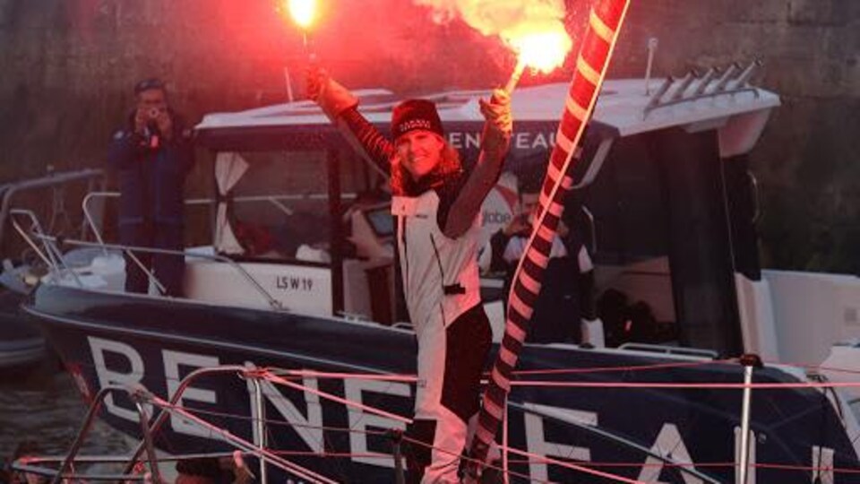 Une femme sourit sur son bateau en tenant deux fumigènes.