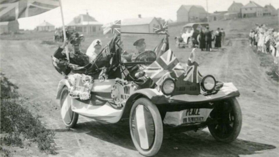 Une vieille voiture d'antan sans toit (1919) avec des soldats et une infirmière à l'intérieur et des drapeaux partout sur la voiture. C'est écrit ''paix'' devant la voiture.
