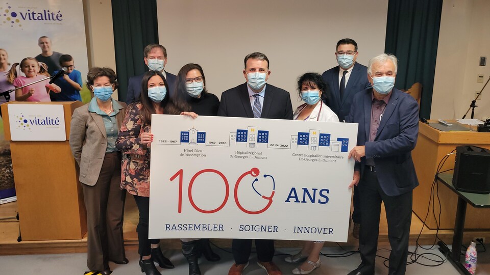 Huit personnes qui portent le masque sanitaire montrent fièrement une pancarte sur laquelle il est écrit : « 100 ans, rassembler, soigner, innover ». Elles sont dans une salle qui ressemble à un amphithéâtre. Un logo du Réseau de santé Vitalité est bien visible derrière elles. 