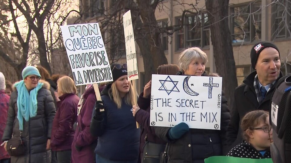Des personnes portent des pancartes sur lesquelles est écrit : « Mon Québec favorise la diversité » et «The secret is in the mix », cette dernière accompagnée de dessins représentant une croix de David, un croissant et une étoile, ainsi qu'un crucifix.