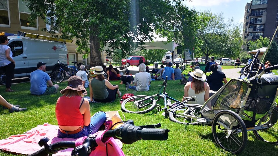 Des gens assis sur l'herbe, dans un parc, regardent vers une scène dressée sous une tente.