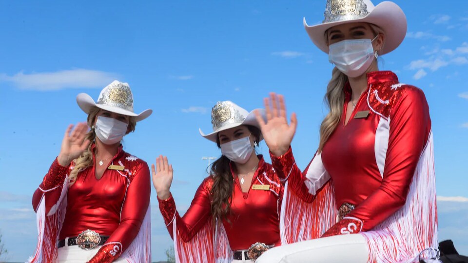 Trois princesses du Stampede saluent la caméra portant leur habit d'apparat et un masque chirurgical lors du déjeuner aux crêpes au volant, le 4 juillet 2020.