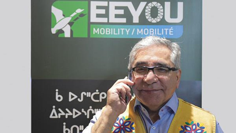Abel Bosum parle au cellulaire devant une affiche d'Eeyou Mobilité.