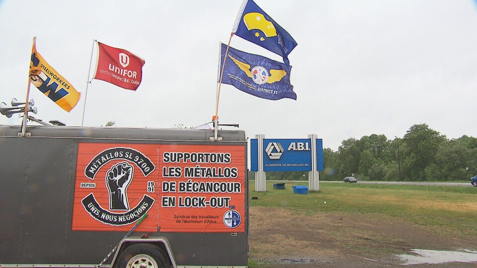 Une insigne où on peut y lire « Supportons les métallos de Bécancour en lock-out » est apposée sur un camion. Plusieurs drapeaux flottent au vent. L'insigne de la compagnie ABI est en arrière-plan.