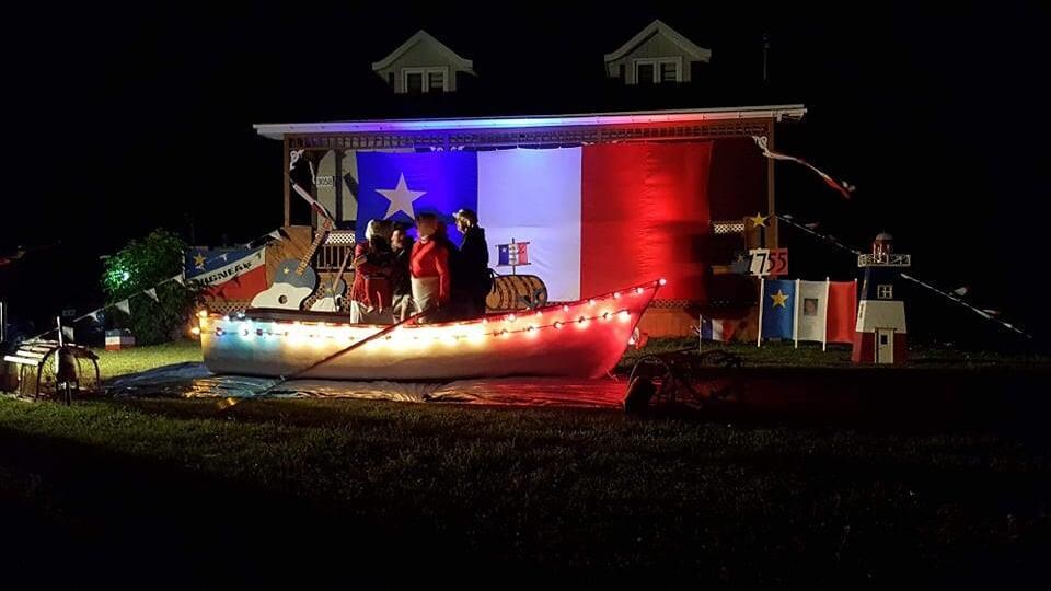 Un bateau installé dans l'herbe devant une maison sur laquelle est tendue un immense drapeau de l'Acadie. 