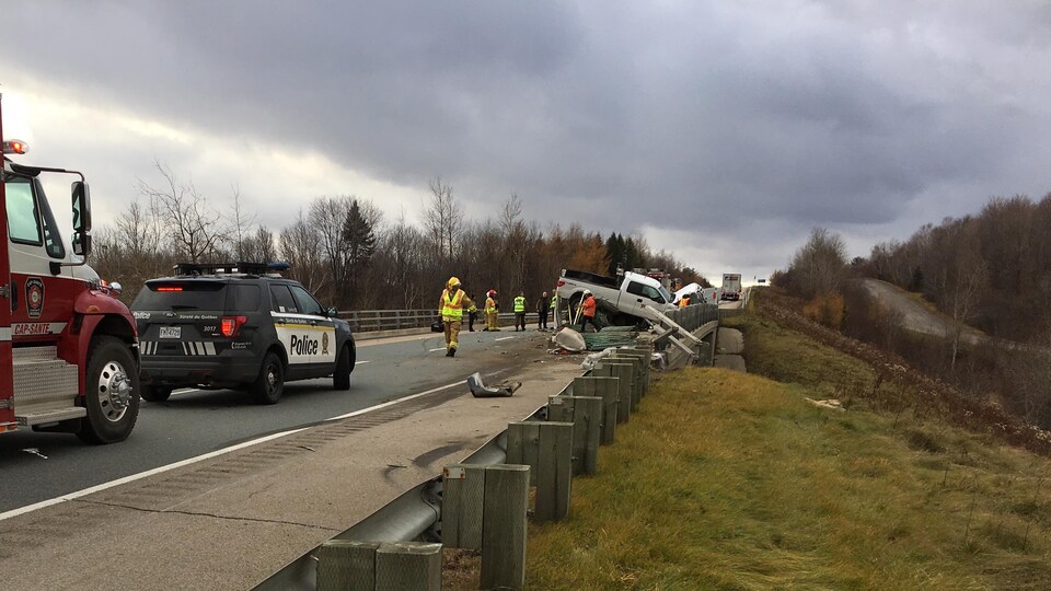 Photo de la scène d’accident. On aperçoit une camionnette blanche accidentée. Un véhicule utilitaire sport de la Sûreté du Québec et un camion de pompier.