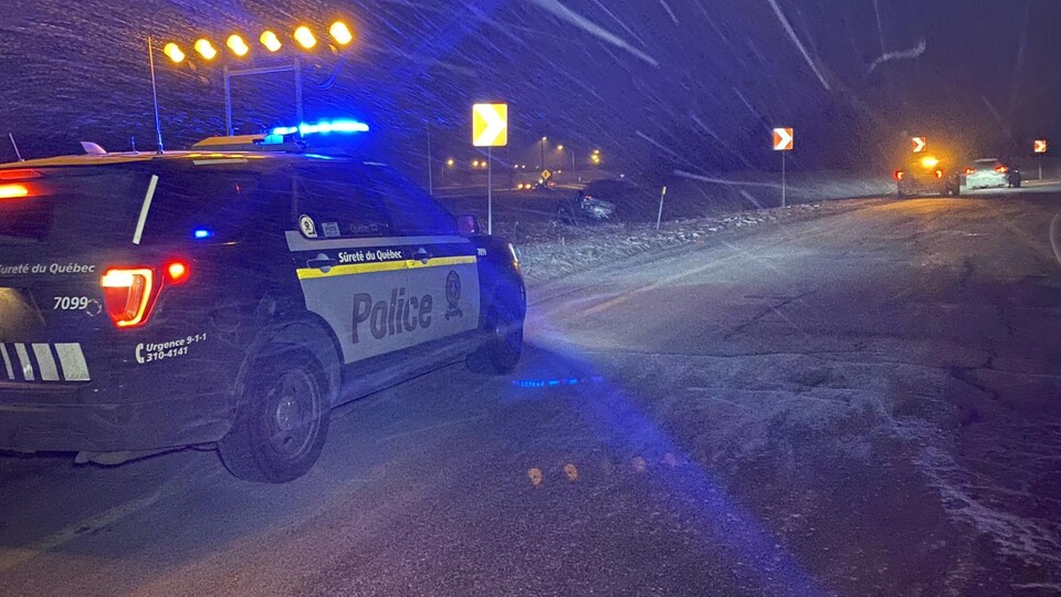 Une voiture de la Sûreté du Québec est stationnée dans la bretelle d'une sortie de l'autoroute 50, avec les gyrophares allumés, en pleine tempête de neige. Quelques mètres plus loin, entre la bretelle et l'autoroute, une voiture est à l'envers dans le banc de neige. 