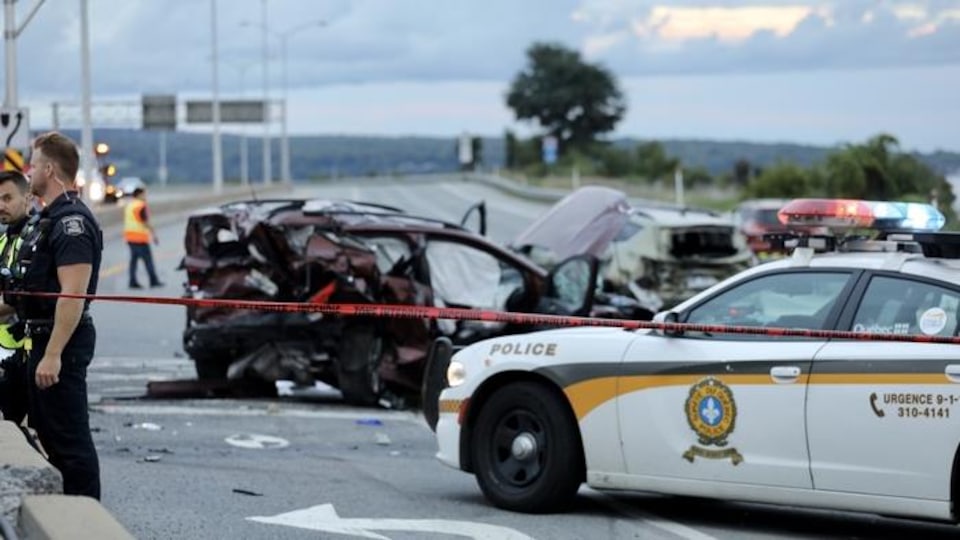 Scène d'un accident de voiture. On voit une voiture complètement cabossée et cassée de partout, une autopatrouille de police et des policiers en uniforme dans un périmètre délimité par un ruban interdisant le passage. Des débris jonchent le sol.