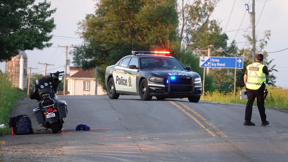 Un policier portant un dossard jaune observe la route au travers de laquelle est stationnée son automobile. La motocyclette accidentée est à sa gauche.       