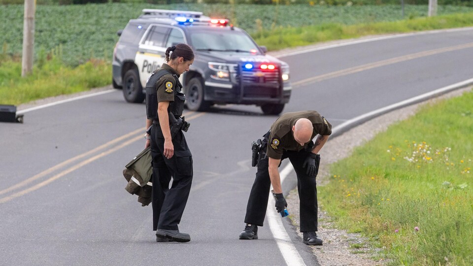 Deux policiers en uniforme sur une route de campagne déserte. L'un d'eux est penché en avant. Une voiture de police est garée au milieu du chemin.
