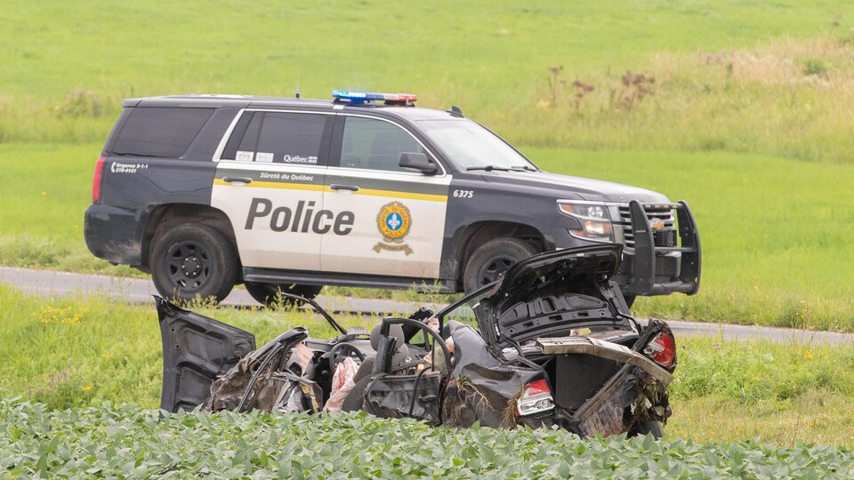 Une carcasse de voiture noire dans un fossé. Une voiture de police est stationnée sur la route.