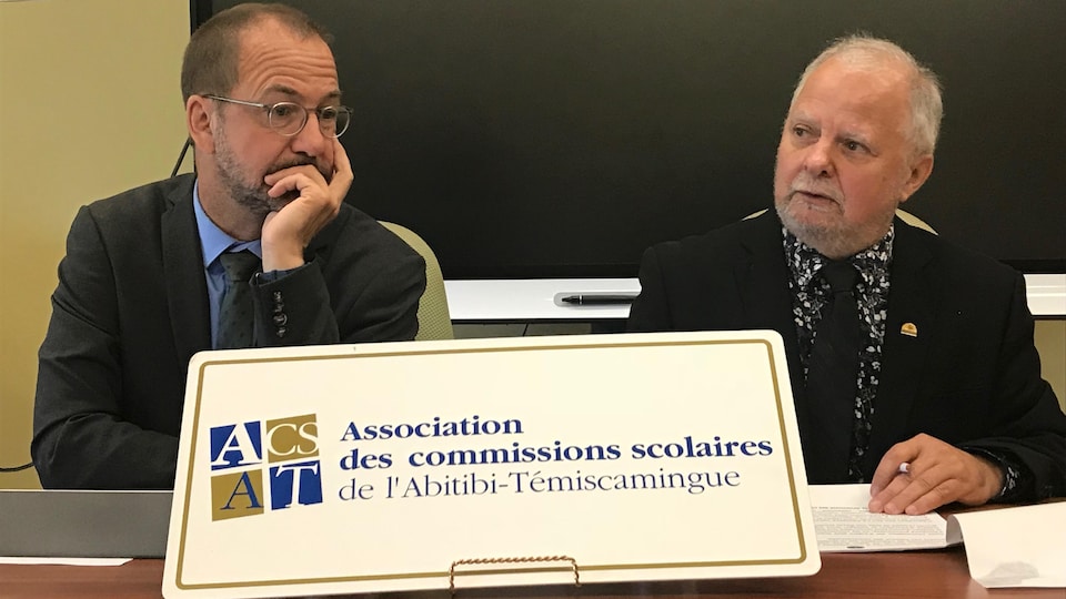 Deux hommes parlent lors d'une conférence de presse derrière une affiche de l'Association des commissions scolaires de l'Abitibi-Témiscamingue.