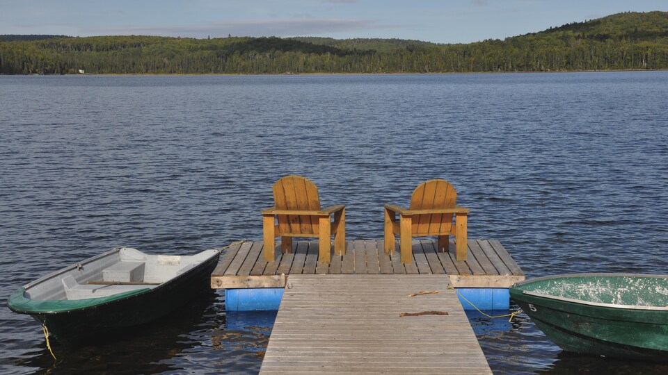 Deux chaises Adirondack se trouvent sur le quai d’un lac en Haute-Mauricie. Deux chaloupes flottent de chaque côté.
